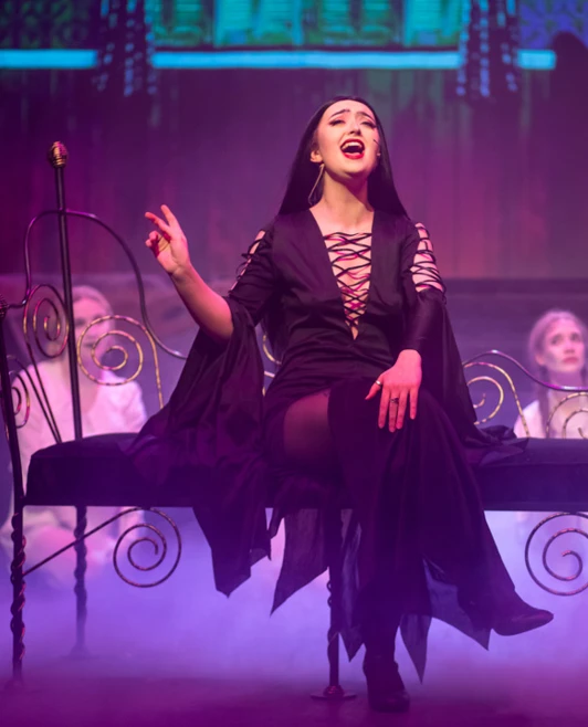 Actress portraying Morticia Addams singing passionately on a gothic-style bed surrounded by ghostly characters Actress portraying Morticia Addams singing passionately on a gothic-style bed surrounded by ghostly characters